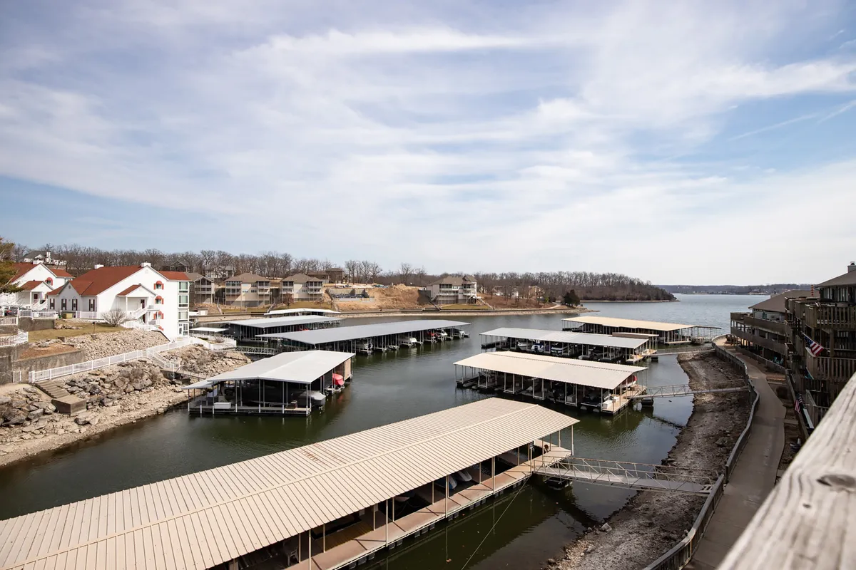 A panoramic view of a marina with boats docked on calm water, surrounded by buildings that reflect the lakeside community atmosphere. The scene captures both boating activity and tranquil moments by the water, with the sunrise or sunset casting a warm glow.