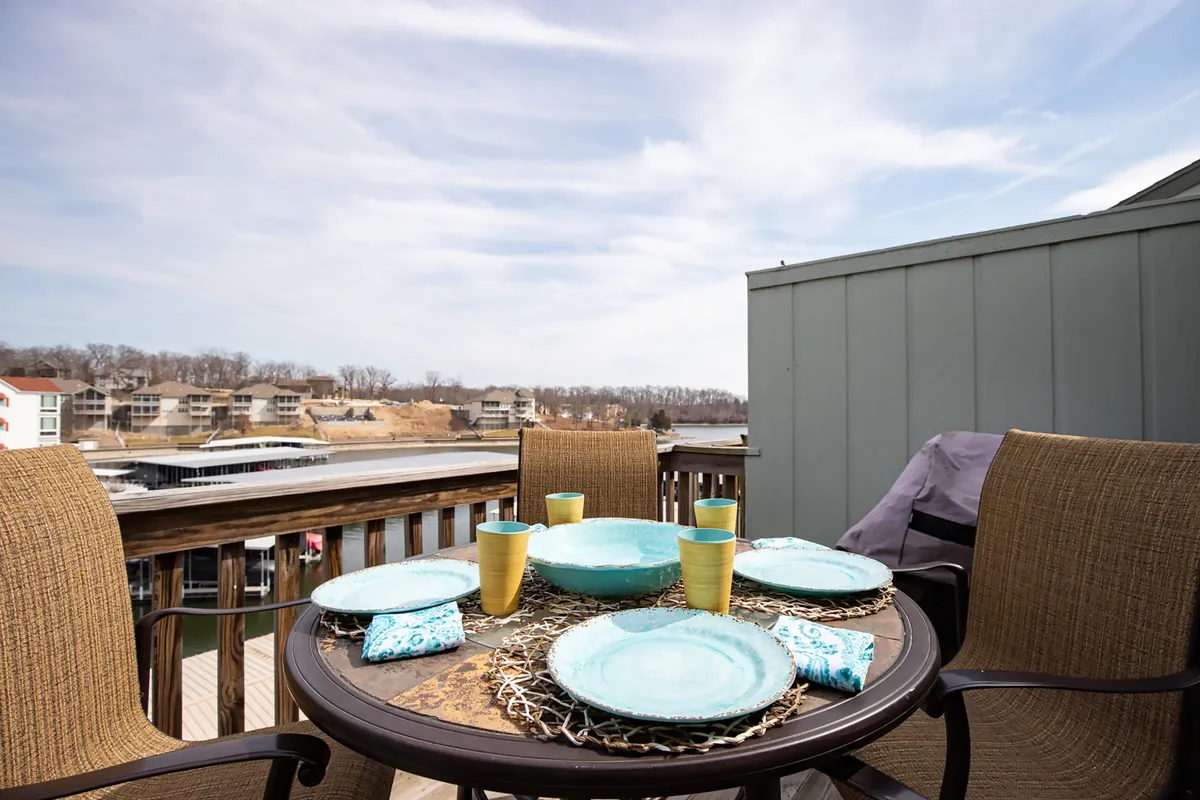 An exterior shot of an outdoor dining table set with blue plates and yellow cups, positioned on a balcony or patio with a sweeping view of the lake in the background. The atmosphere is bright and cheerful, evoking the enjoyment of meals and socializing outdoors while taking in the scenery.