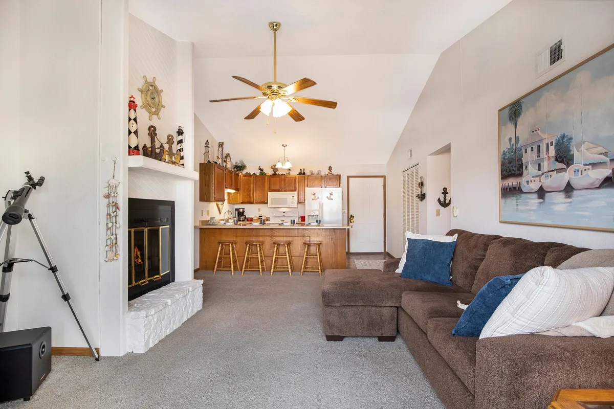 An expansive open-plan living space with a comfortable couch, kitchen island with bar stools, and a fireplace. Large windows fill the area with sunlight, creating a bright backdrop for family gatherings or unwinding after a day at the lake. The layout highlights the home's social and relaxing vibe.