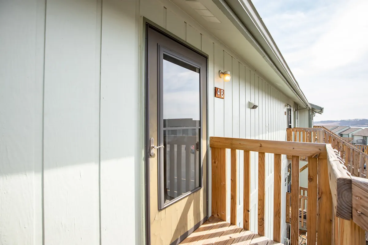 A welcoming exterior view of a condo entrance with a wooden railing, the front door labeled '4B', and space for relaxing or socializing on the balcony. The natural light and open layout evoke a sense of arrival and the possibility of outdoor gatherings.
