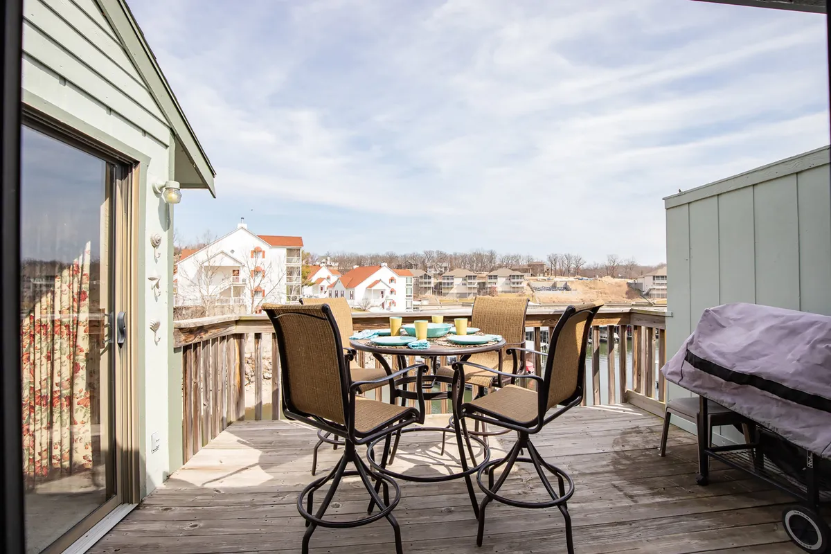 The image showcases an inviting round dining table with chairs arranged on a private patio or balcony, overlooking the sparkling lake and nearby buildings. The tranquil water view is front and center, with space for guests to relax, share meals, or enjoy coffee while watching boats drift by.