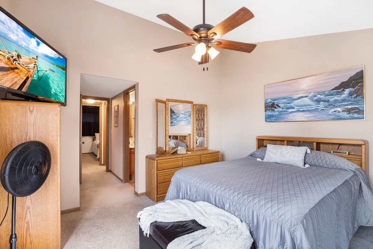 A cozy bedroom with a gray quilted bed, a dresser with a mounted TV, a large mirror, and a landscape painting. The space is designed for comfort, with ample storage and relaxing touches that make it welcoming for guests of all ages.