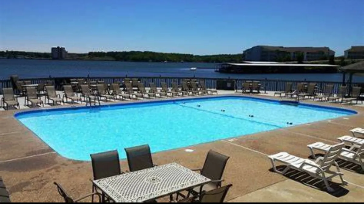 This exterior image features a sparkling outdoor swimming pool surrounded by lounge chairs, with beautiful lake views in the background. The scene is active with guests swimming, sunbathing, and socializing, capturing the leisurely resort atmosphere. Sunset lighting adds a warm, inviting glow, highlighting the proximity of the pool to the lake.