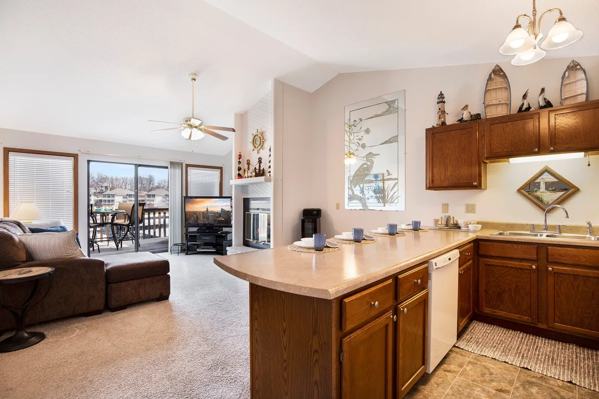A cozy seating area adjacent to a kitchen counter, featuring a fireplace and large windows opening to the balcony and the lake beyond. The room is bathed in natural light, with inviting decor and comfortable furniture that encourages guests to unwind and enjoy the setting.