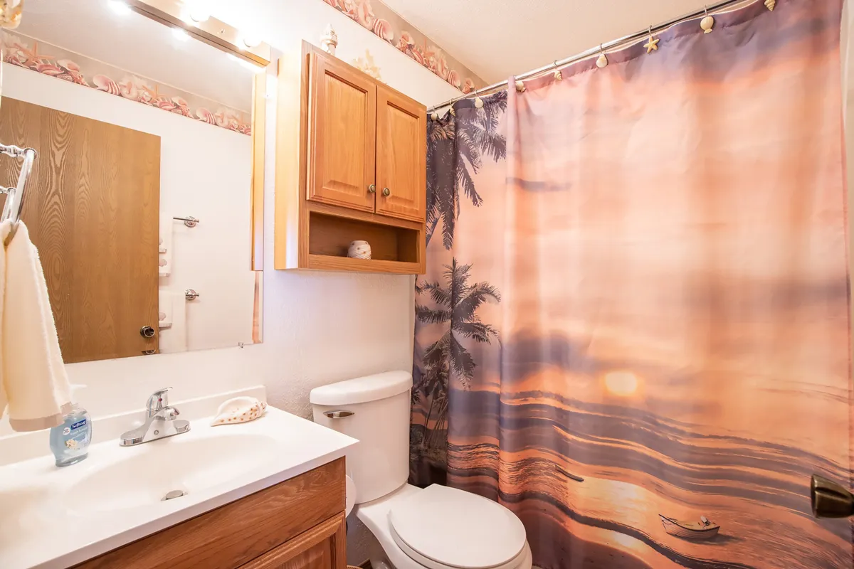 A tranquil bathroom featuring a vibrant shower curtain with a sunset and palm tree motif, wooden cabinetry, and a bright sink area. The space feels inviting and well-equipped for guest comfort, offering a fresh start or relaxing end to the day.