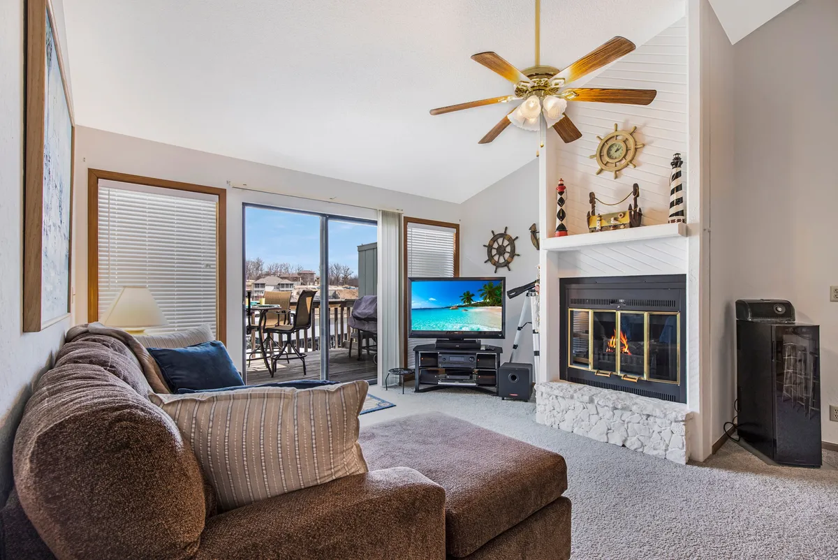 A cozy living room featuring a central fireplace (for display), a plush sofa, a sliding glass door letting in ample daylight, and a TV. The furnishings and layout create a warm, welcoming atmosphere ideal for relaxing, watching movies, or entertaining guests. The sliding door and windows emphasize the indoor-outdoor connection and hint at scenic views.