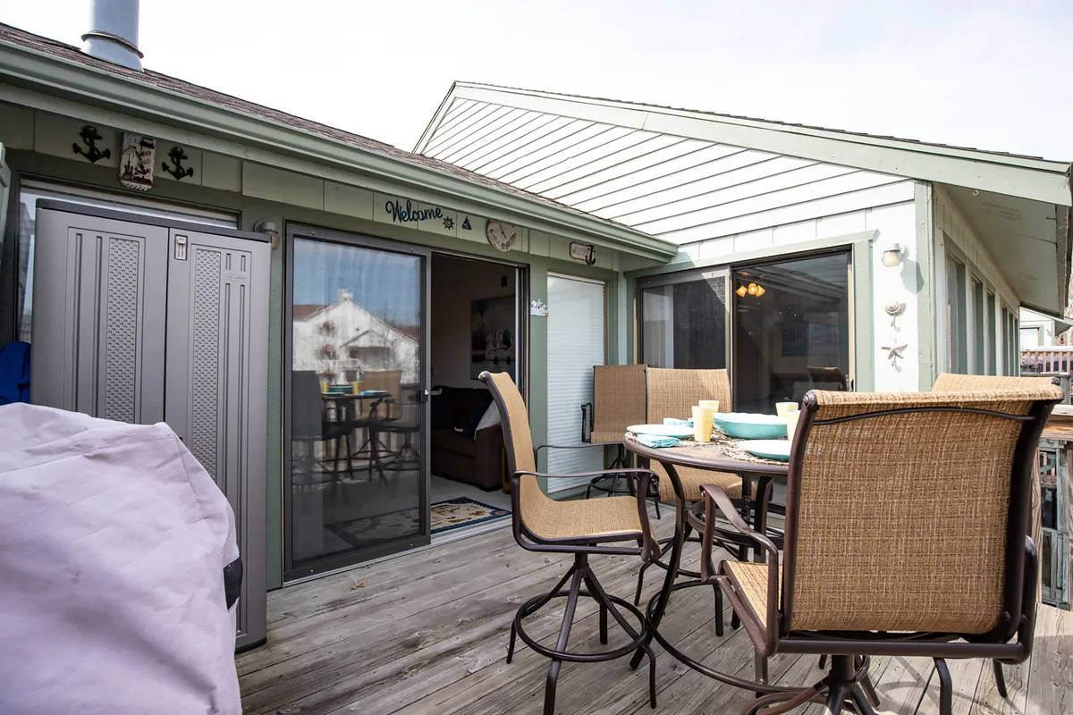 An exterior patio scene featuring a dining table set for outdoor meals, comfortable outdoor seating, decorative wall elements, and doors leading inside. The atmosphere is welcoming and set for sunset dinners or morning coffee, with a focus on social gatherings and relaxation.